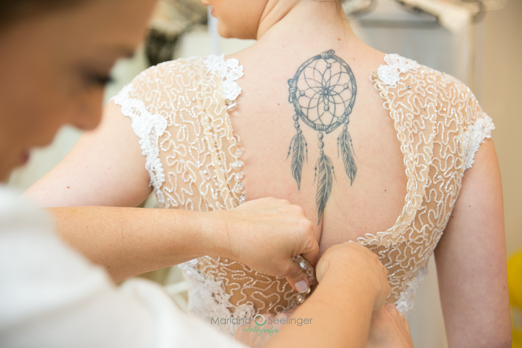 Madrinha do casamento fechando o vestido da noiva em foto q mostra a tatuagem da noiva  em fotografia de Mariana Seelinger Fotógrafa de Casamentos em Niterói e RJ