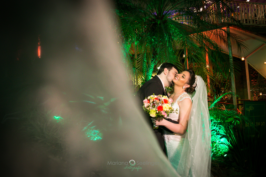 Foto quem quee mostra o véu da noiva e ao fundo os noivos abraçados em fotografia de Mariana Seelinger Fotógrafa de Casamentos em Niterói e RJ