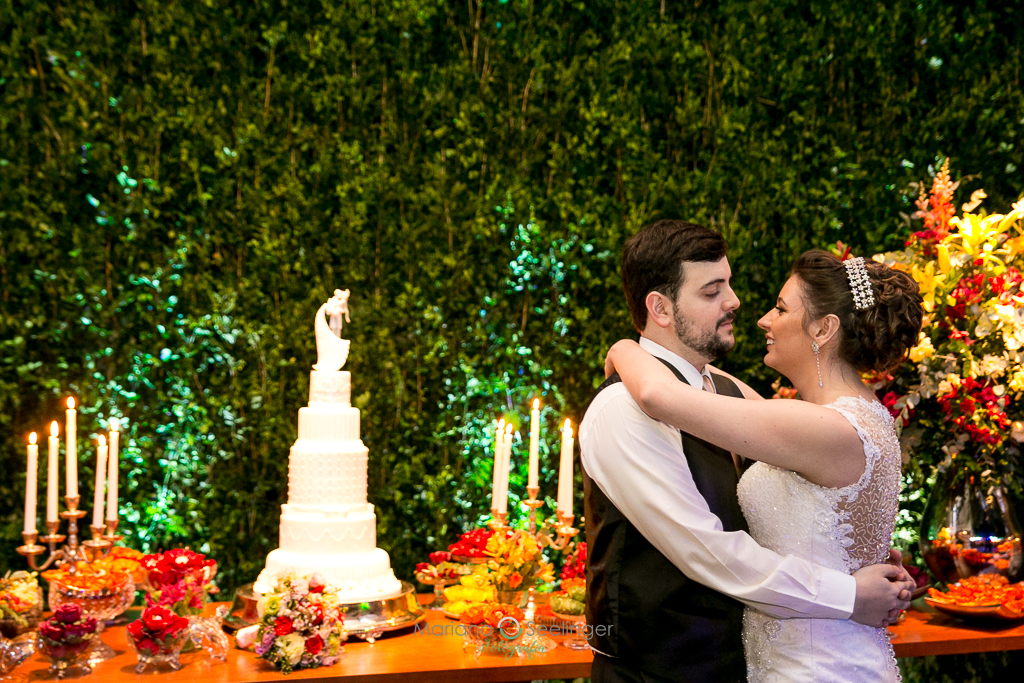 Foto do casal de noivos em frente a mesa do bolo de 5 andares branco com bonecos em cima em fotografia de Mariana Seelinger Fotógrafa de Casamentos em Niterói e RJ