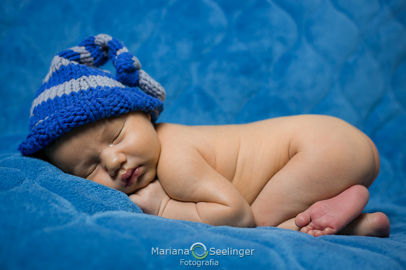 bebê deitado em acolchoado azul com touquinha de tricô em Niterói - RJ