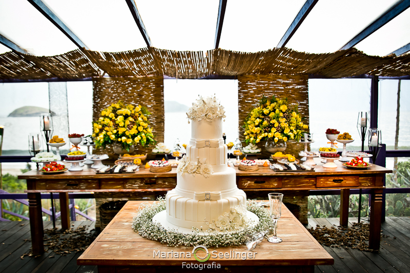 bolo de 4 andares apoiado em mesa de madeira rústica com decorações de flores amarelas ao fundo em fotografia de Mariana Seelinger Fotógrafa de Casamentos em Niterói e RJ