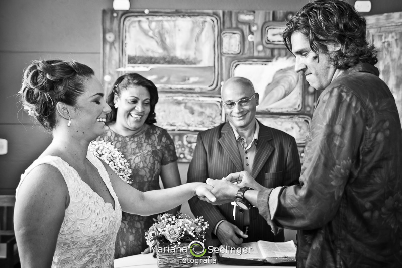 Noivo colocando aliança no dedo de sua noiva com o celebrante ao fundo em fotografia de Mariana Seelinger Fotógrafa de Casamentos em Niterói e RJ