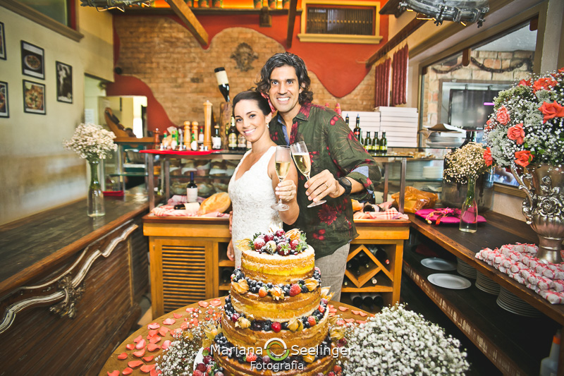 Noivos felizes brindando próximos ao seu bolo bastante colorido em fotografia de Mariana Seelinger Fotógrafa de Casamentos em Niterói e RJ