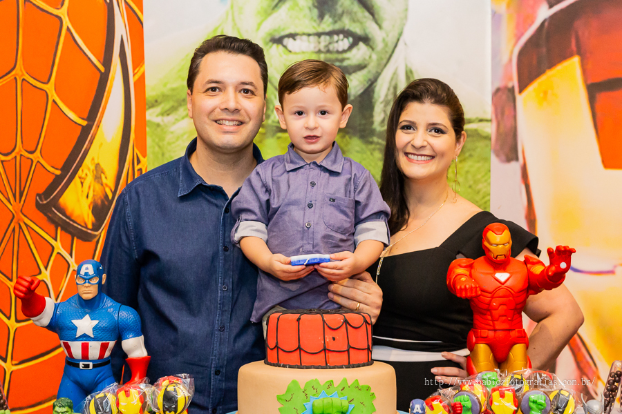 Fotografia do aniversariante Theo com os pais na mesa do bolo, com decoração da liga da justiça e vingadores no Jungle Frends em Maringá.