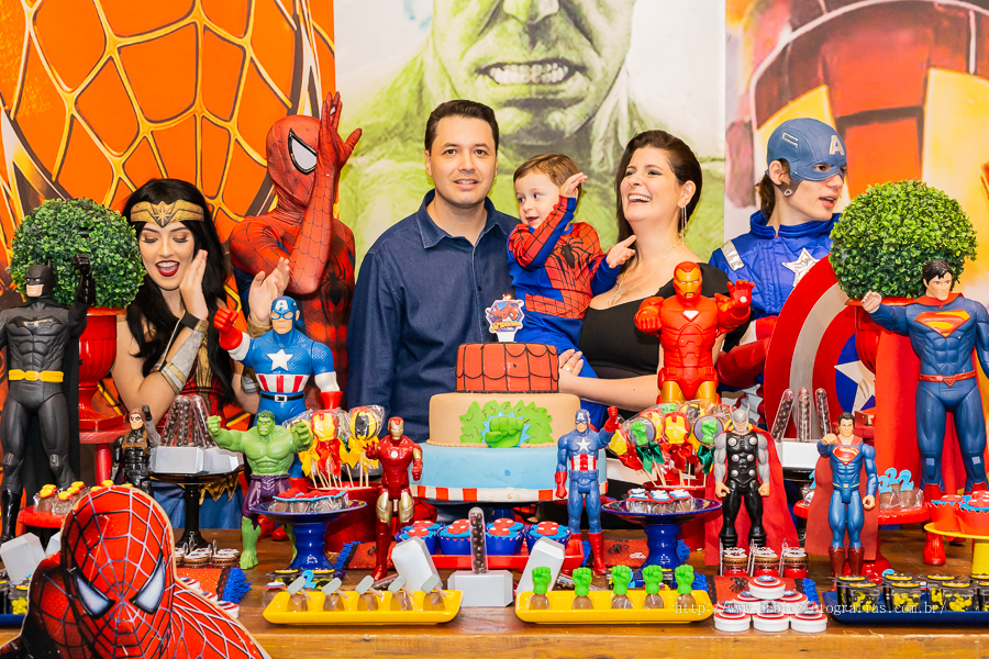 Foto do aniversariante Théo com os pais na mesa do bolo cantando parabens com os personagens da liga da justiça e vingadores no buffet Jungle Frends em Maringá.