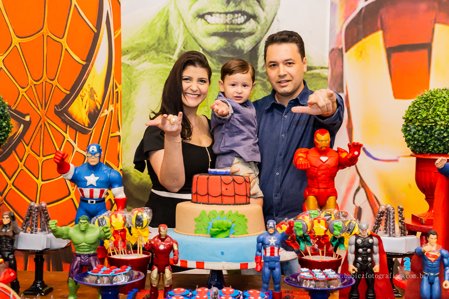 Fotografia do aniversariante Theo com os pais na mesa do bolo, com decoração da liga da justiça e vingadores no buffet Jungle Frends em Maringá.