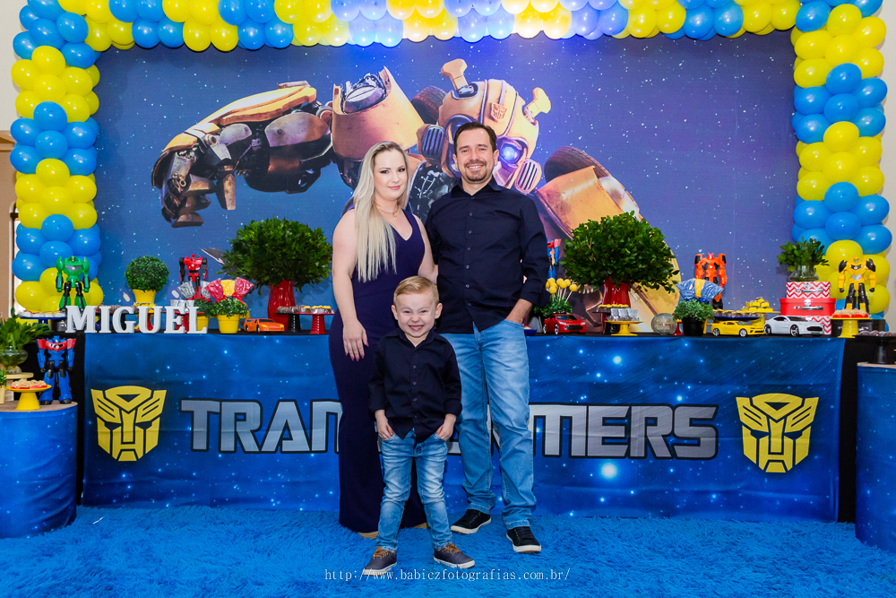fotografia do aniversariante com os pais em frente a mesa de bolo da decoração transformes