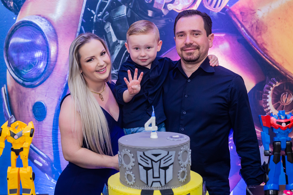 fotografia do aniversariante com os pais em frente a mesa de bolo da decoração transformes