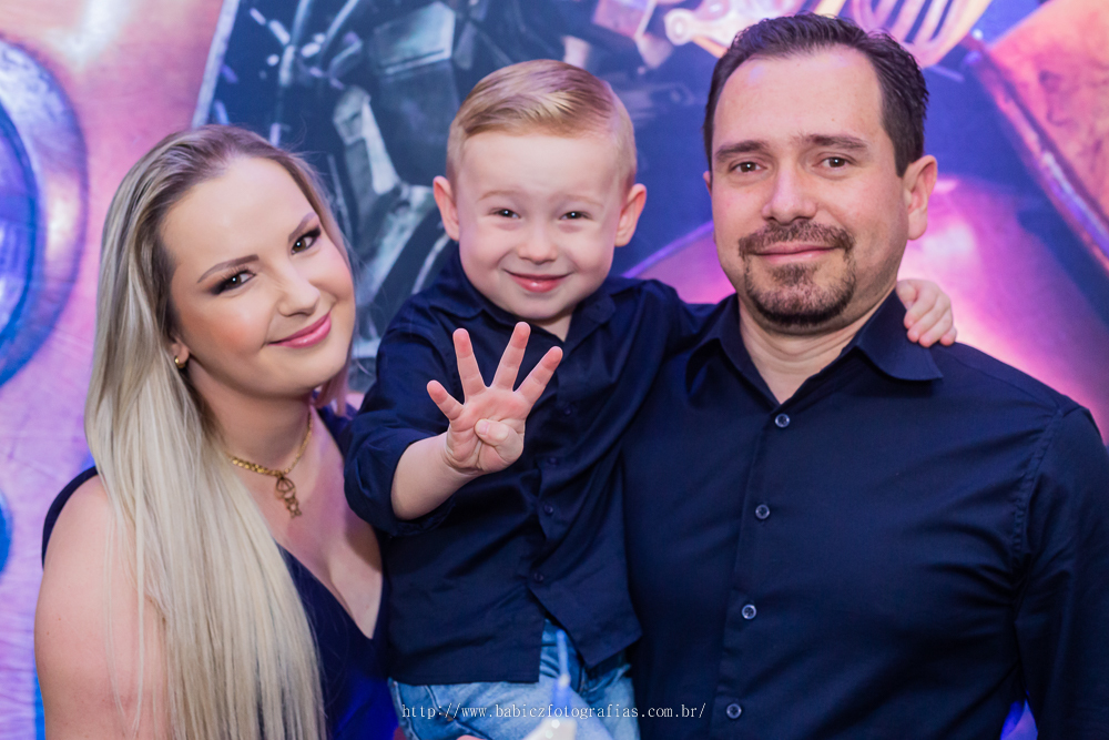 fotografia do aniversariante com os pais em frente a mesa de bolo da decoração transformes