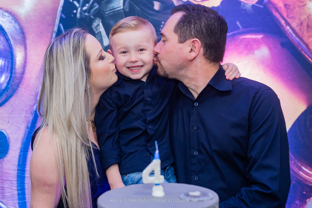fotografia do aniversariante com os pais em frente a mesa de bolo da decoração transformes