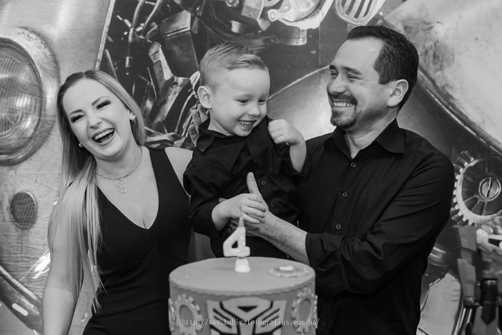 fotografia do aniversariante com os pais em frente a mesa de bolo da decoração transformes em preto e branco