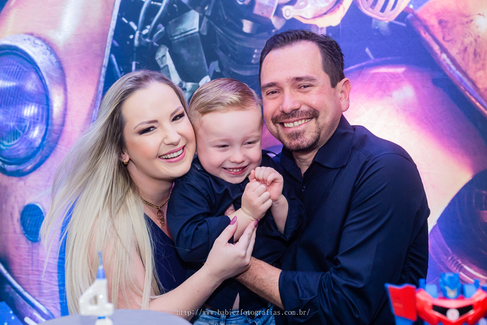 fotografia do aniversariante com os pais em frente a mesa de bolo da decoração transformes