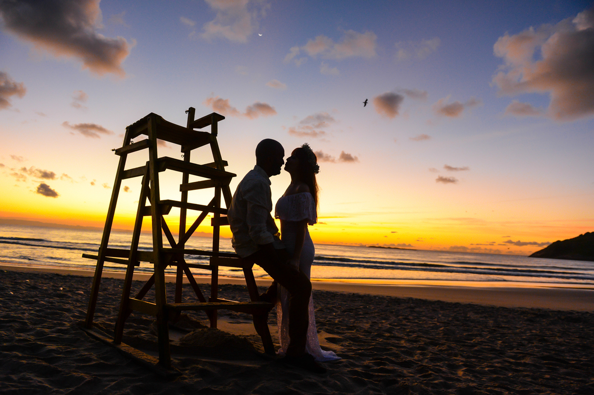 foto do casal somente silueta  sentado na escada do salva vidas  escada de frente um para o outro  bem juntos com o sol nascendo  no mar ao fundo 