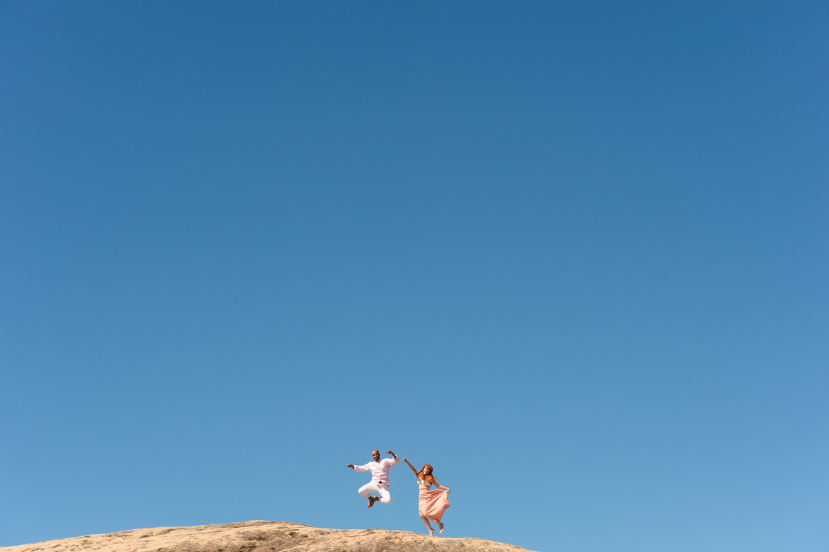aquela foto minimalista feita na praia com o casal pulando em cima de uma pedra enorme com o ceu azul de fundo 