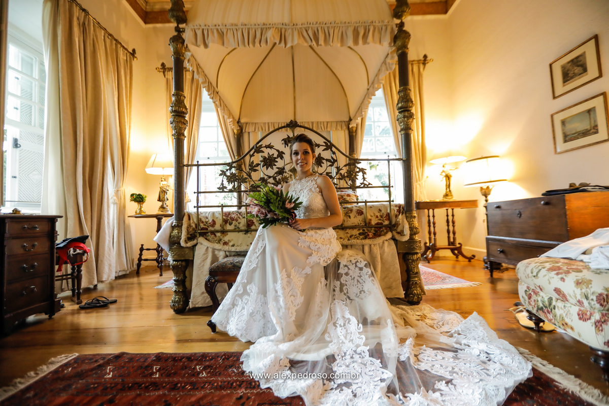 noiva sentada com vestido todo bordado com a calda aberta com uma cama antiga de fundo e com buque nas maos