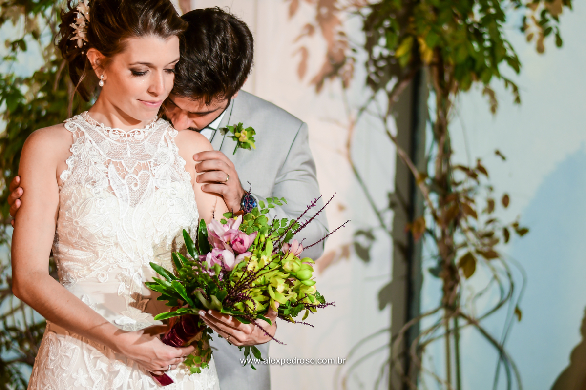 noiva com o buque de flores na mao e o noivo abraçando ela por tras beijando seu ombro