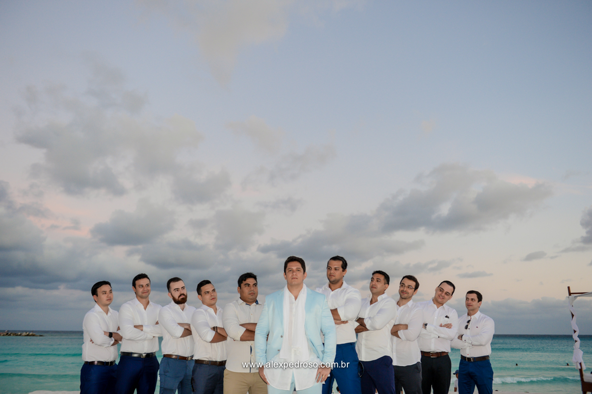 noivo no centro e suas padrinhos  ao lado com o mar e o ceu azul ao fundo 