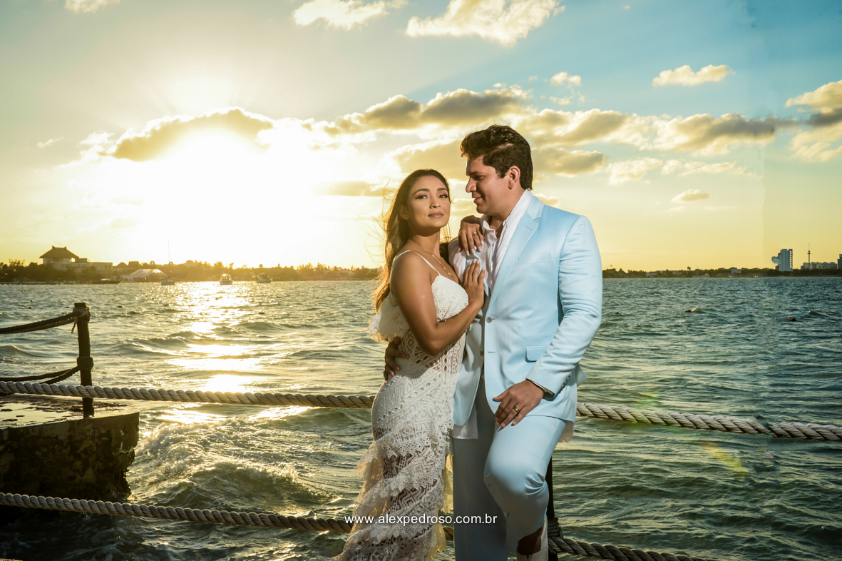 noiva vestindo brando com um vestido mais solto e noivo vestindo terno Azul Clora do Ricardo almeida  bem juntos com o por do sol ao fundo o mar aparecendo no deck no mar do caribe no mexico 