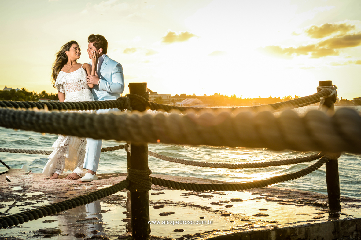 noiva vestindo brando com um vestido mais solto e noivo vestindo terno Azul Clora do Ricardo almeida  bem juntos com o por do sol ao fundo o mar aparecendo no deck no mar do caribe no mexico 