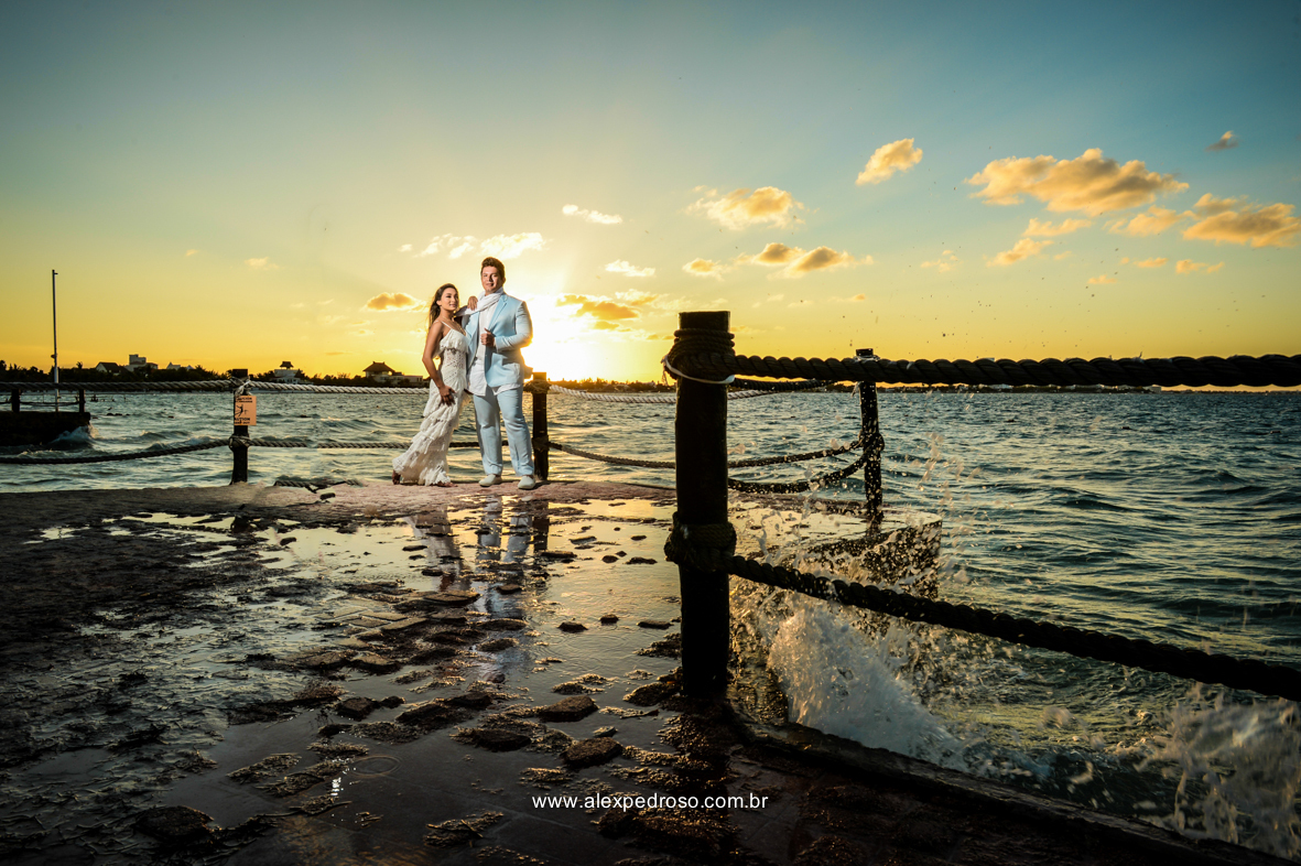 noiva vestindo brando com um vestido mais solto e noivo vestindo terno Azul Clora do Ricardo almeida  bem juntos com o por do sol ao fundo o mar aparecendo no deck no mar do caribe no mexico 