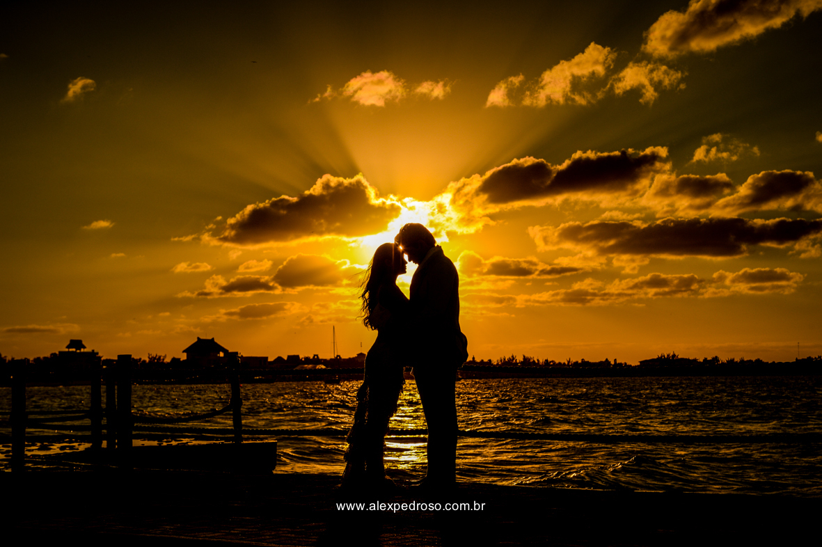 noiva vestindo brando com um vestido mais solto e noivo vestindo terno Azul Clora do Ricardo almeida  bem juntos com o por do sol ao fundo o mar aparecendo no deck no mar do caribe no mexico 