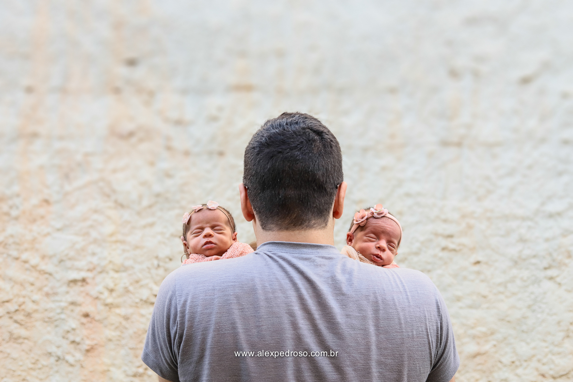 Pai segurando dois bebês de costas pra câmera, mostrando só os rostos de suas filhas dormindo, usando uma camisa cinza, parede de fundo.