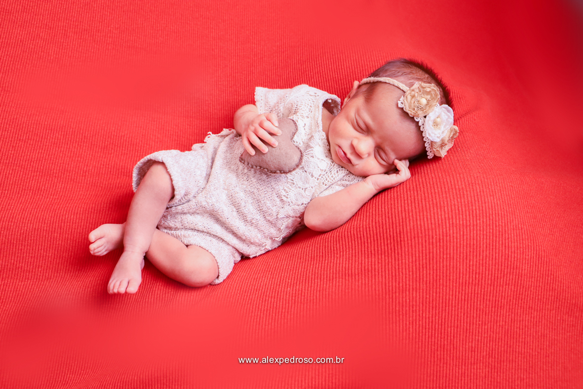 Bebê  dormindo com um fundo vermelho, segurando um coração de pelúcia apoiado no peito com a mão no rosto,  usando um macacão branco  e uma tiara de flores.