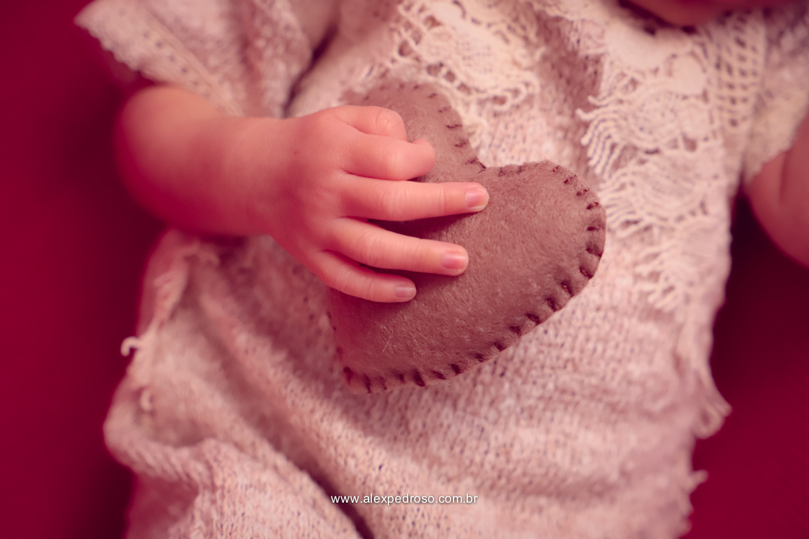Detalhes de um neném segurando um coração de pelúcia e sua mãozinha pequena, apoiados no peito, usando um macacão branco.