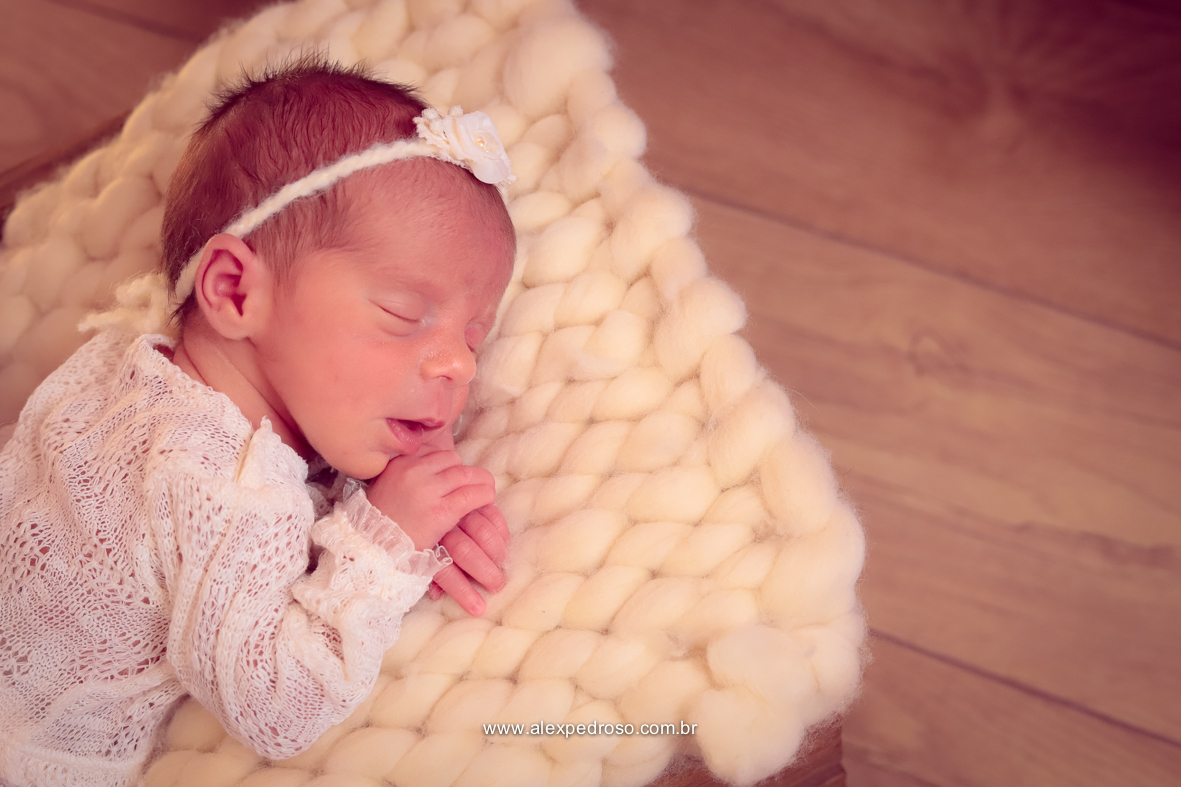 Bebê dormindo apoiado sobre as duas mãos, usando uma roupa branca e uma tiara branca de flor, sobre um cobertor/manta bordada.