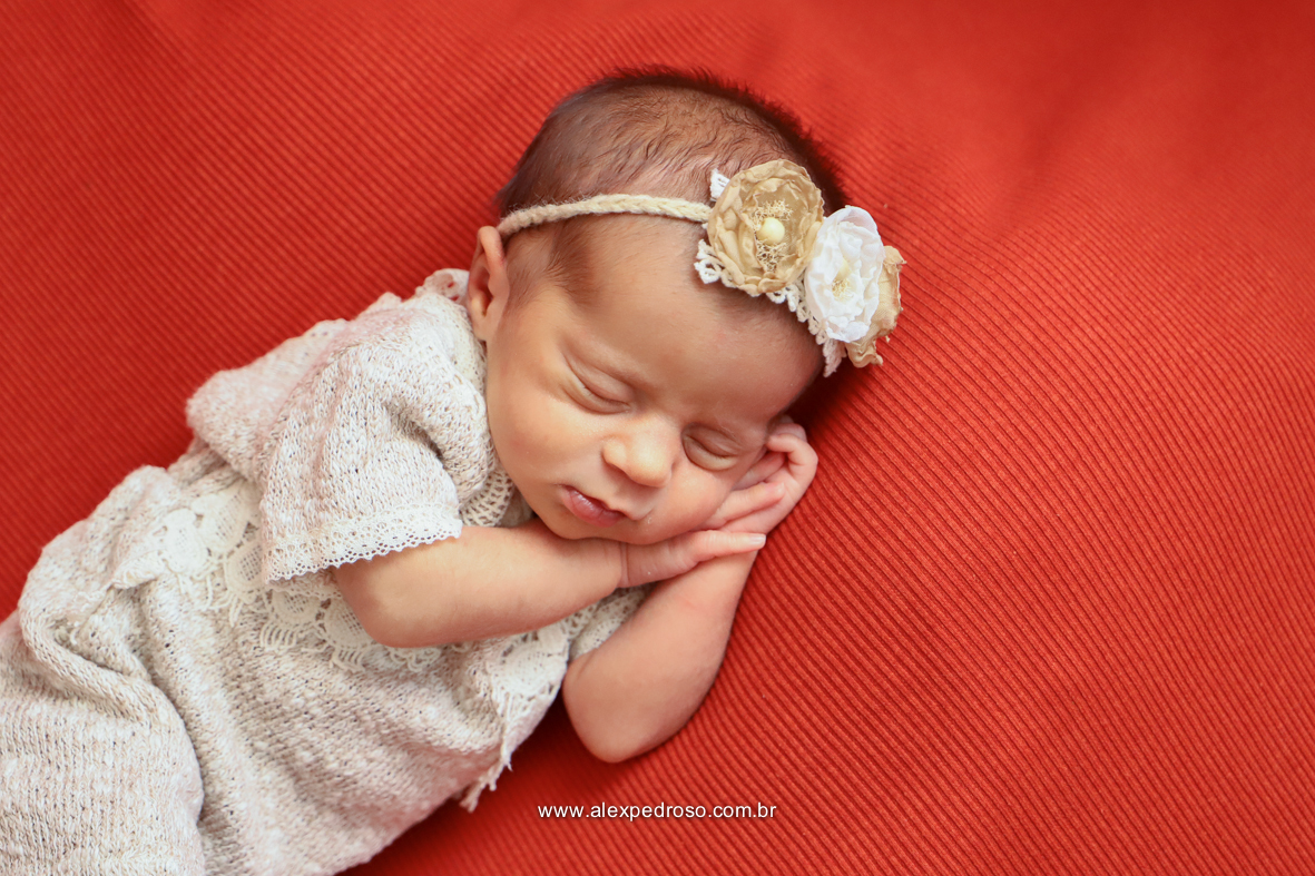 Bebê dormindo apoiado sobre as duas mãos, usando um macacão branco e uma tiara de flores em um fundo vermelho.