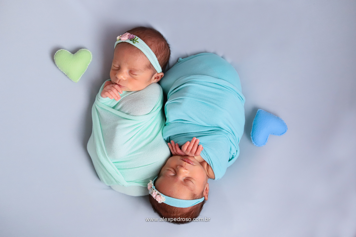 Dois bebês dormindo enrolados no wrap um verde água e um azul claro, com faixas de suas respectivas cores e com corações do lado de cada um com a mesma cor.