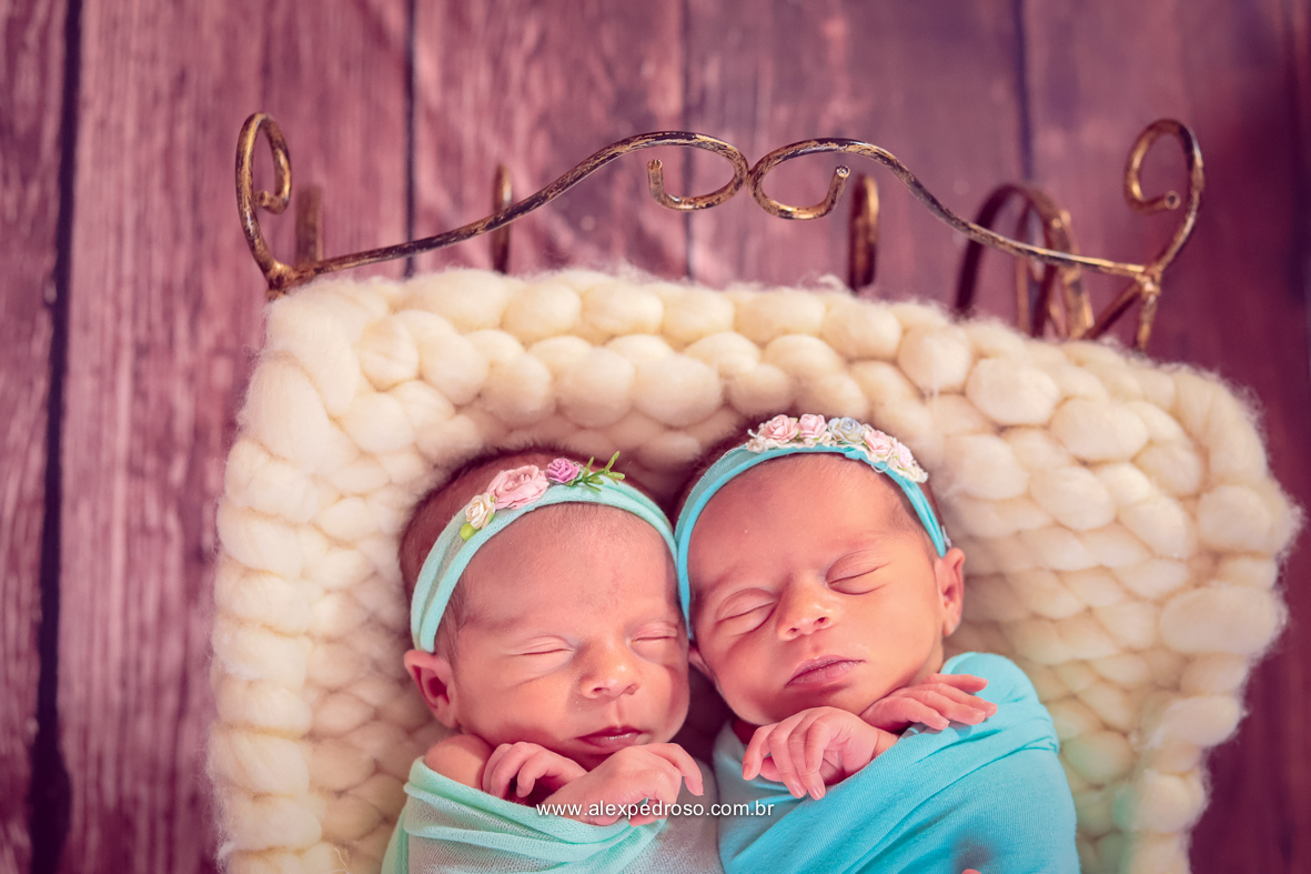 Dois bebês dormindo enrolados no wrap um verde água e um azul claro, sobre uma manta bordada em um berço de ferro, com o piso de madeira de fundo.