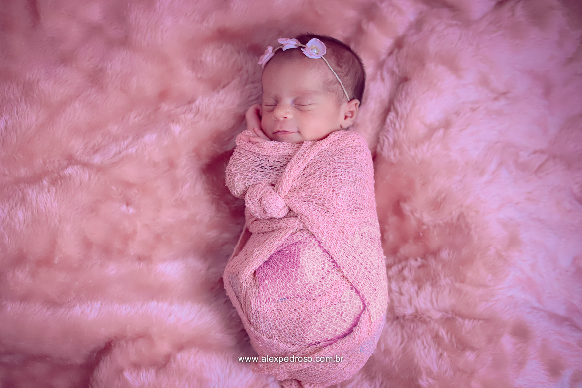 Bebê dormindo e sorrindo enrolado no wrap rosa, sobre um cobertor da mesma cor e uma faixa também.