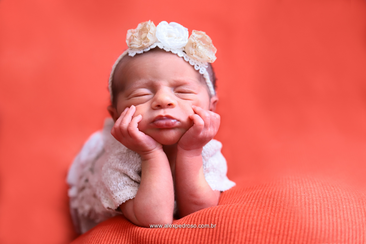 Bebê dormindo de barriga pra baixo apoiado sobre suas duas mãos, usando um macacão e uma tiara de flor sobre um fundo vermelho.