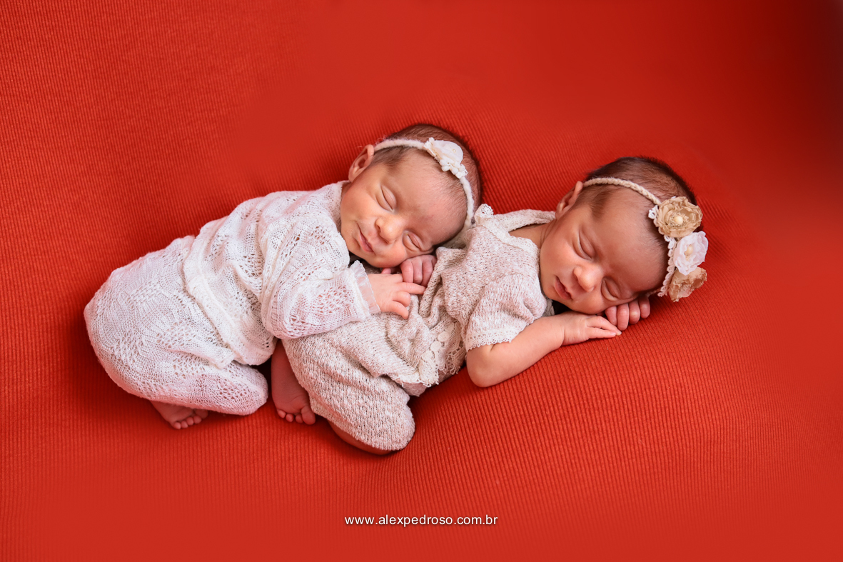 Bebês dormindo uma sobre a outra, usando roupas brancas e uma tiara de flor sobre um fundo vermelho.
