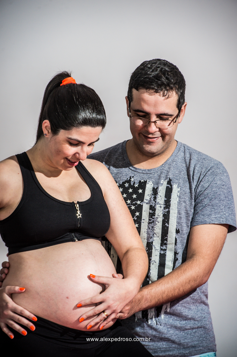 Casal de gestante, futuro pai e mãe olhando sorrindo para a barriga e com as mãos nela, gestante com um top preto e esposo com uma camiseta cinza, foto pegando apenas da metade da barriga pra cima