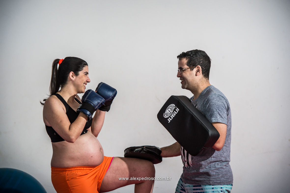 Casal de gestante treinando boxe em uma academia, mulher usando um top preto e um short laranja com as luvas de boxes azul e com o joelho esquerdo levantado e o futuro pai com um aparador de chute treinando juntamente com a sua esposa