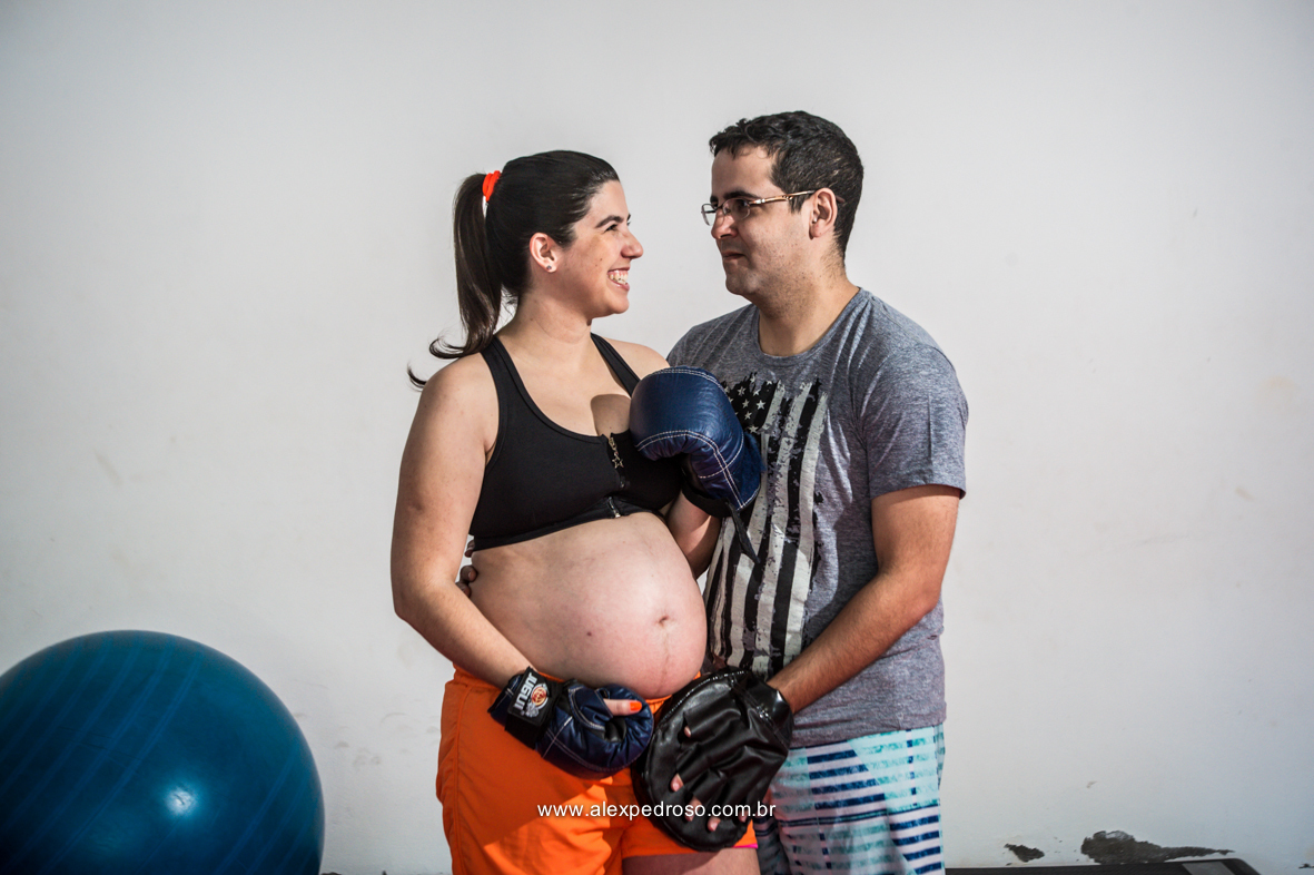 Casal de gestante em uma academia, mulher com luvas de boxe azul segurando a barriga com um top preto e um short laranja e seu esposo ao lado com a mão na barriga também tendo equipamento de boxe nas mãos, e uma bola azul no fundo das foto, 