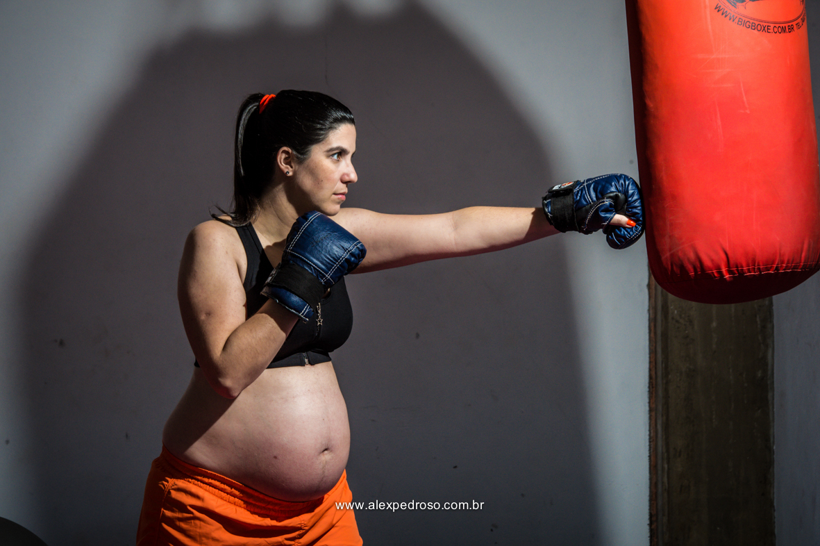 Gestante praticando boxe com as luvas azuis em um saco de pancada vermelho em uma academia, com um top preto e um short laranja, com a sombra do saco de boxe por trás