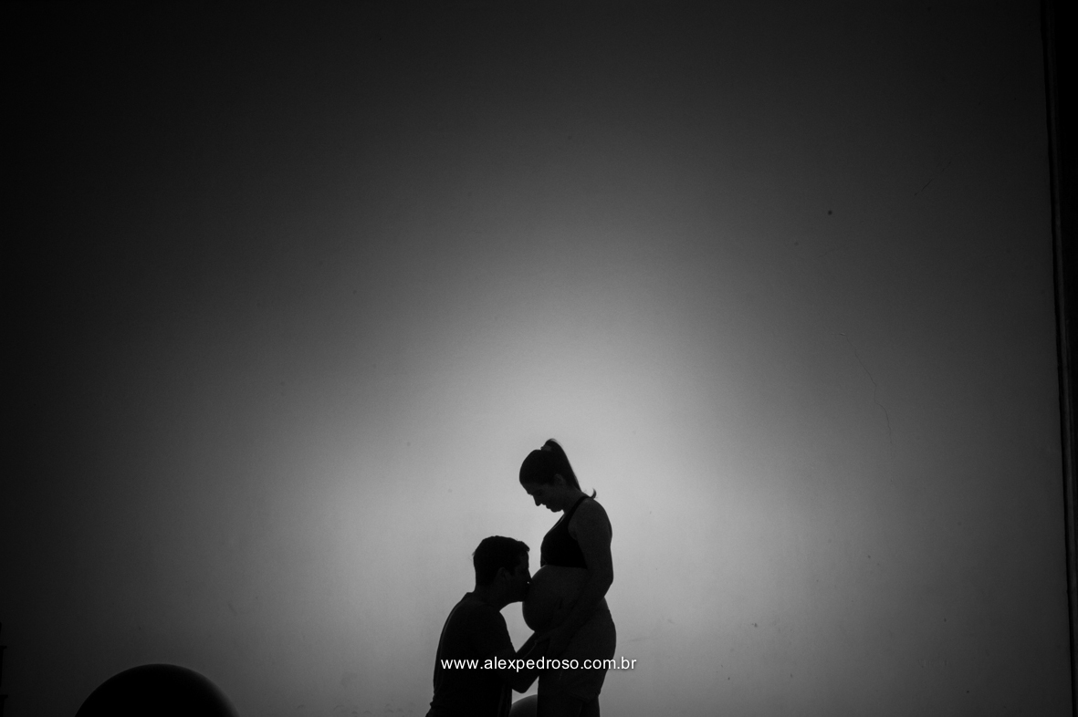 Futuro pai ajoelhado beijando a barriga da mãe gestante, em uma foto preta e branca e a mãe olhando  para o pai, os dois com a mão na barriga da gestante