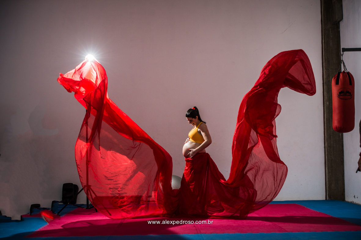 Foto de gestante em uma academia com tecidos vermelhos jogados pra cima, formando uma borboleta com um top laranja, tendo um saco de pancada vermelho ao lado