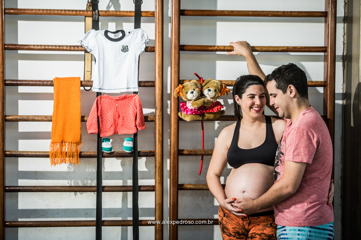 Casal de gestante, onde os dois seguram a barriga com o neném, tendo cabeças encostadas e os dois sorrindo, gestante apoiada de costas para parede e o futuro pai de lado encostado com a mão na parede, foto em academia tendo em voltas equipamentos e roupas