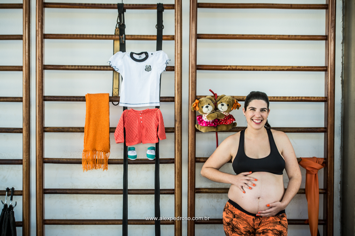 Mãe gestante com a mão na barriga sorrindo encostada em uma parede feita de madeiras, com roupas da criança penduradas juntamente com o sapatinho e o ursinho de pelúcia, foto realizada em academia