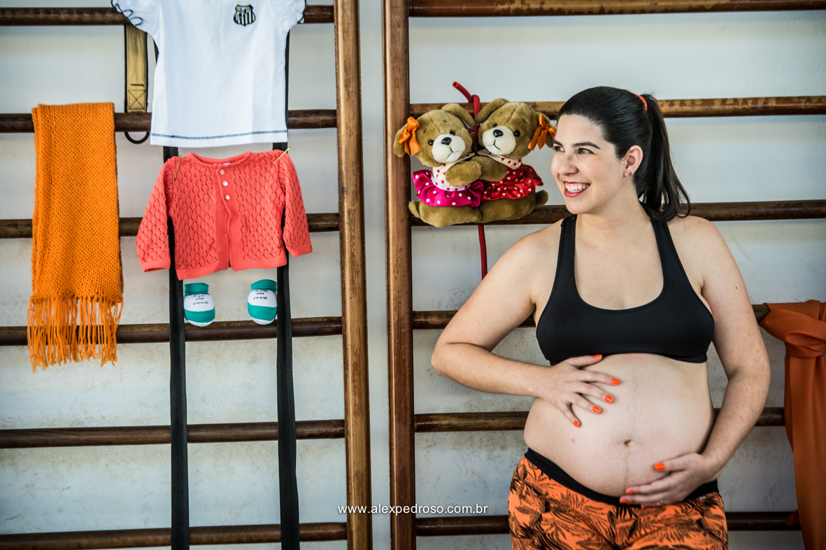 Mãe gestante com a mão na barriga sorrindo encostada em uma parede feita de madeiras, com roupas da criança penduradas juntamente com o sapatinho e o ursinho de pelúcia, foto realizada em academia