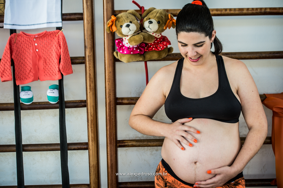 Mãe gestante com a mão na barriga sorrindo encostada em uma parede feita de madeiras, com roupas da criança penduradas juntamente com o sapatinho e o ursinho de pelúcia, foto realizada em academia da barriga pra cima