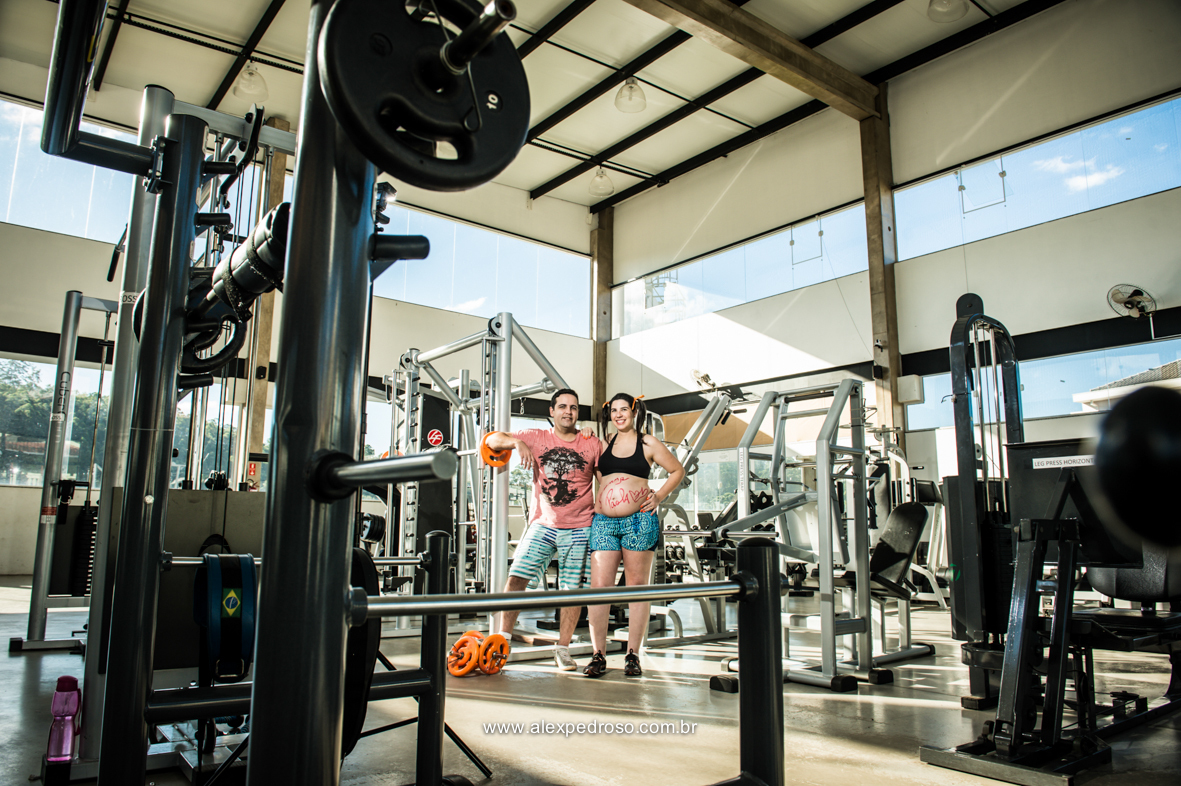 Casal com a mãe gestante em uma parte da academia tendo em volta equipamentos para treinar, futura mãe com a mão na cintura e futuro pai apoiado em uns dos equipamentos