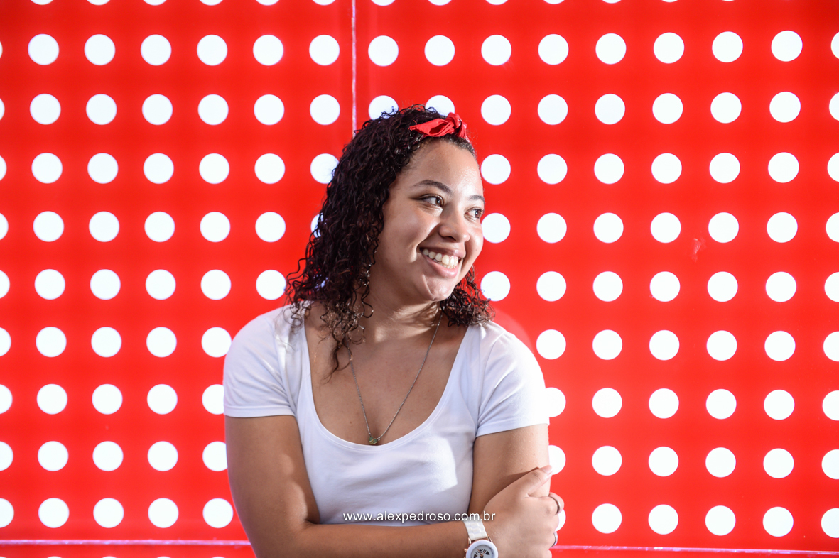 Menina de cabelo cacheado em um fundo vermelho com bolinhas brancas, sorrindo olhando para o lado com a mão no outro braço e um relógio branco, usando uma blusa branca e uma bandana vermelha, com um escapulário, foto da barriga pra cima,tirada na paulista