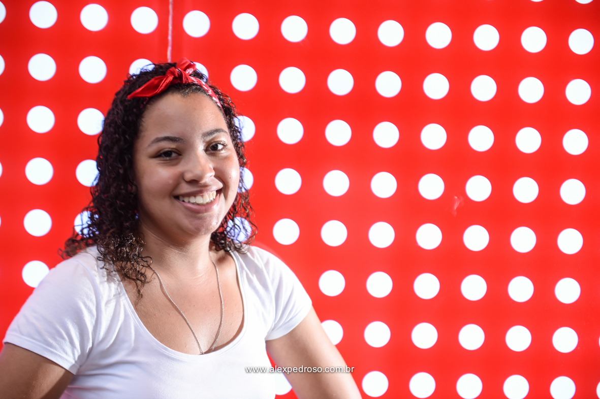 Menina de cabelo cacheado em um fundo vermelho com bolinhas brancas, sorrindo, usando uma blusa branca e uma bandana vermelha, com um escapulário no pescoço, foto tirada na paulista.