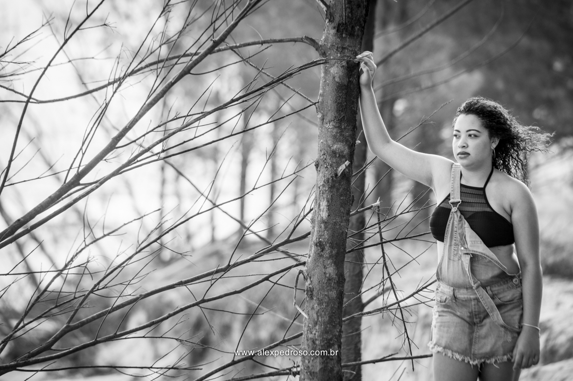 Menina de cabelo cacheado apoiada em uma árvore, olhando para o horizonte usando um cropped preto e um macacão jeans, foto em preto e branco tirada em uma praia, do joelho pra cima.