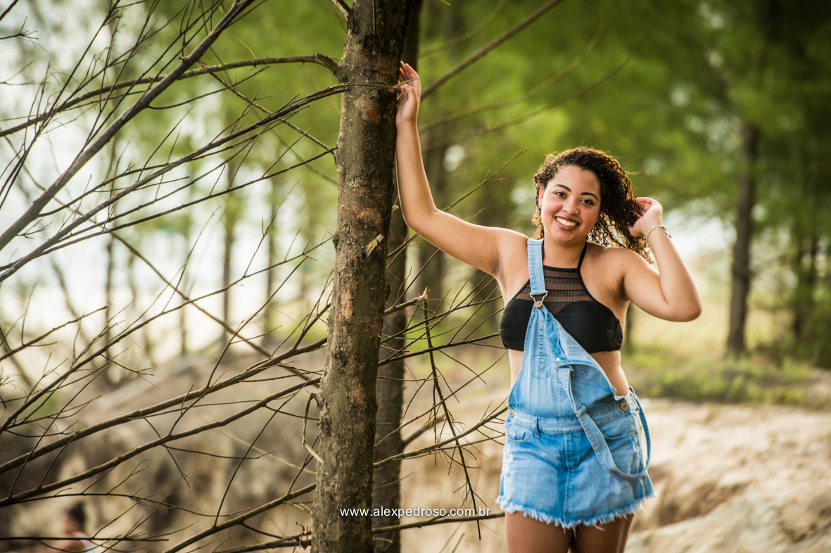 Menina de cabelo cacheado mexendo em seu próprio cabelo com uma mão e a outra apoiada em uma árvore, sorrindo, usando um cropped preto e um macacão jeans, foto tirada em uma praia, do joelho pra cima.